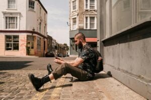 bearded man sitting on the sidewalk and taking photos of the street with his smartphone